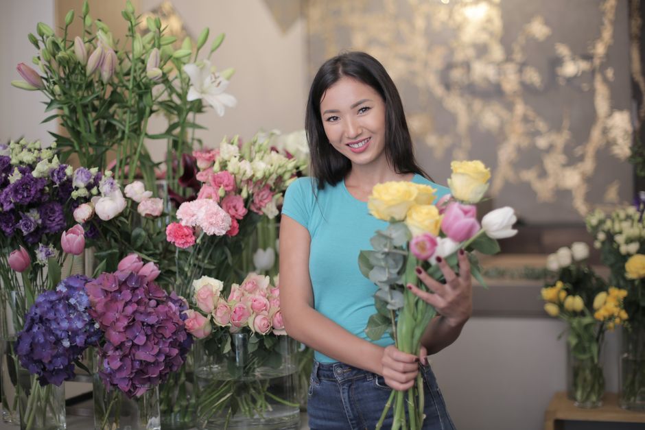 A young woman with shoulder-length dark hair and a friendly smile, wearing a light blue T-shirt, is holding a bouquet of fresh flowers including yellow roses, pink tulips, and white eustoma, wrapped i