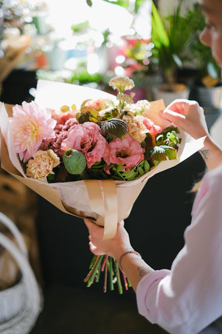 A person holding a beautifully arranged bouquet of fresh flowers indoors, featuring large pink peonies, white and purple blossoms, and lush green foliage. The bouquet is wrapped in white paper and tie
